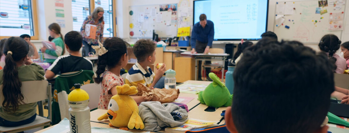 Schulkinder sitzen in einem Klassenzimmer und hören dem Lehrer vorne zu.