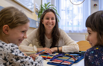 Ehrenamtliche sitzt mit zwei Kindern am Tisch und begleitet sie beim Malen und Schreiben